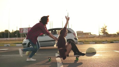 Side view of a woman with her hands raised up sitting on a longboard while her friend is pushing her behind and running during sunset. Enjoying life. Lens flare. Slowmotion shot