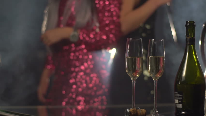 Dancing girls in the sparkling dresses at the background are taking two glasses of champagne.