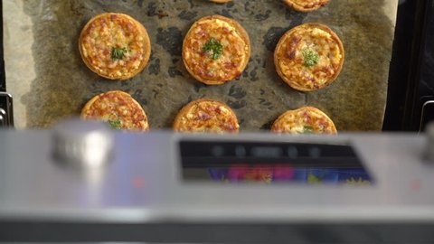 Cooking mini pizza topped with mozzarella cheese and ham in the oven. View from above of chef hands takes out baking sheet with baked small pizzas from the oven.