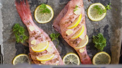 Cooking whole roasted fish with lemon and herbs baked in the oven. View from above of chef hands places baking sheet with raw ocean perch into the hot oven.