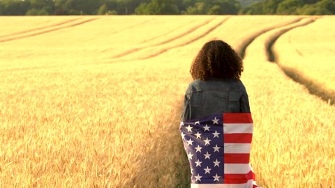 4K video clip of mixed race African American girl teenager female young woman holding an American USA Stars and Stripes flag in a wheat field at sunset or sunrise Stock Video