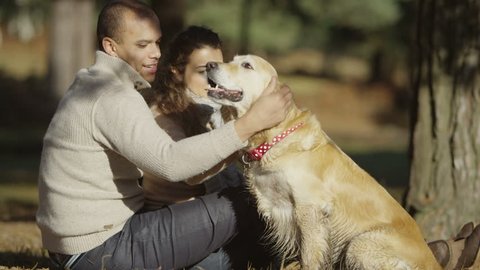 A romantic young couple sit in a close embrace, as they enjoy some leisure time outdoors with their loveable pet dog. 