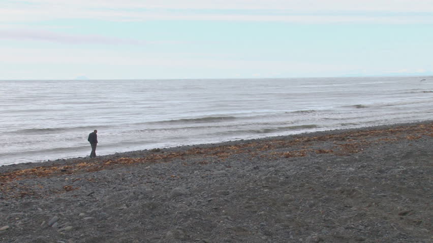 Man walks along beach, backlit by grey and blue skies reflected on moody waters