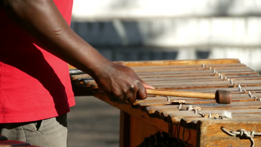 Close up of slight above view of wooden xylophone bars with dark skinned arms & hand rhythmically hitting the bars with out of focus passing people behind. 