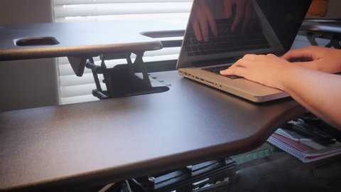 Woman changes the height on a variable standing desk. The work from home mother stays healthy with her ergonomic sit-to-stand office furniture. She lowers the table and continues her online shopping.