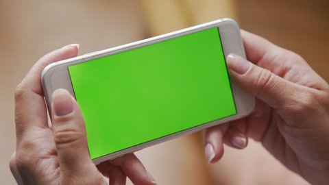 Female hands holding a white smartphone with a green screen on the background of a wooden floor. Modern technologies and gadgets: using the phone. The phone is in a horizontal position.