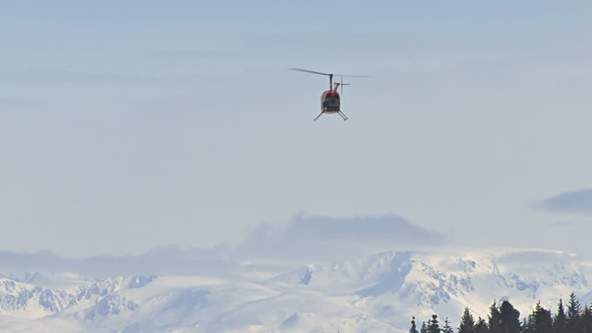 Red 2-man helicopter (Robinson R22) flying away toward Alaskan mountains in the