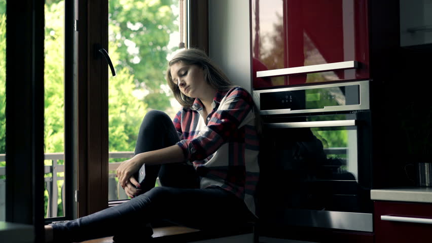 Sad, pensive woman sitting on windowsill in kitchen at home 
