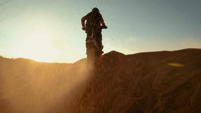 Low Angle Following Shot of the Professional Motocross Motorcycle Rider Driving Over the Dune and Further Down the Off-Road Track. It's Sunset and Track is Covered with Smoke/ Mist.