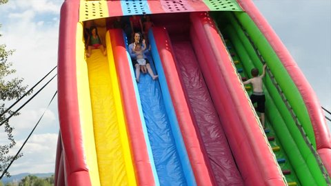Slow motion shot of people going down a large bouncy inflatable slide and almost shooting off the end while at a summertime party