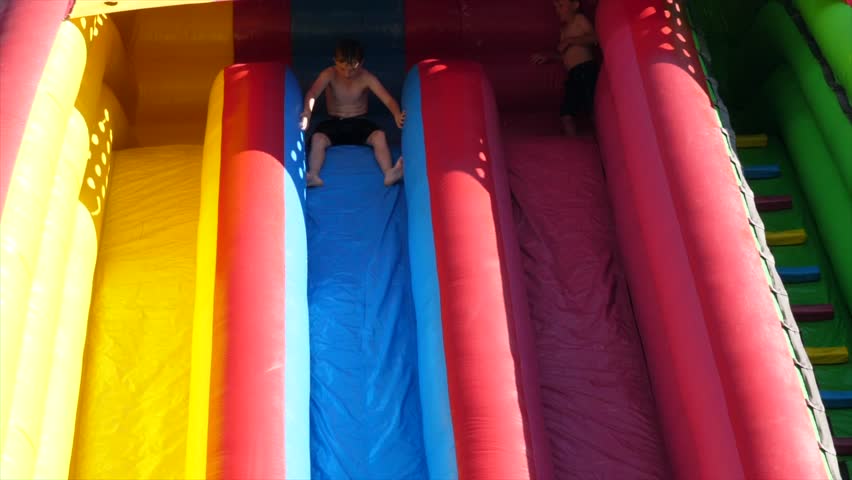 Slow motion shot of people going down a large bouncy inflatable slide and almost shooting off the end while at a summertime party