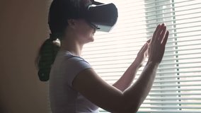 Beautiful young girl stands near the window with virtual reality glasses. Young excited woman wearing VR headset touching objects in virtual world, using goggles mobile application smartphone