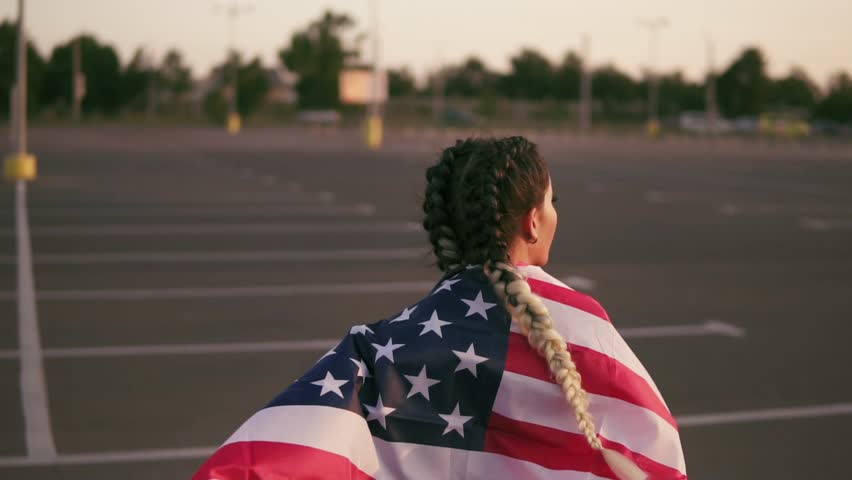 Proud american girl holding stars and stripes. She wraps the american flag over her shoulders and walks forward. Back view. Slowmotion shot