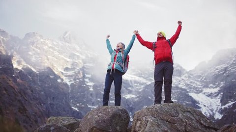 Hikers Reaching Mountain Top. SLOW MOTION. Young happy hiking couple cheering, reached the summit. Man and Woman trekking with backpacks in snow capped mountains. Success and achievement concept