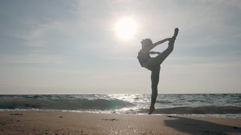 Young woman in casual style - denim and black top doing ballet at the beach. Attractive ballerina practices in stretching on sandy coastline in autumn. Slow motion.