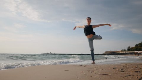 Young woman in casual style - denim and black top doing ballet at the beach. Attractive ballerina practices in stretching on sandy coastline in autumn. Slow motion.