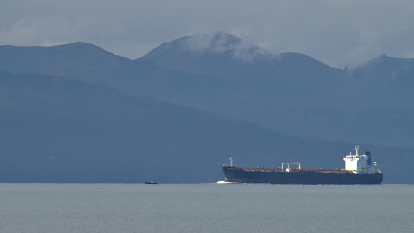 Oil tanker cruising slowly into the bay, passing a smaller boat. Mountains and