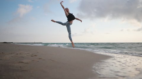 Young woman in casual style - jeans and black top doing ballet at the beach. Attractive ballerina practices in jumping on sandy coastline in autumn. Slow motion.