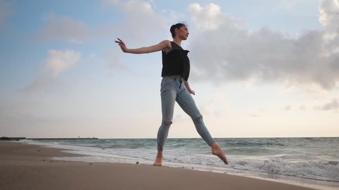 Young woman in casual style - denim and black top doing ballet at the beach. Attractive ballerina practices in jumping on sandy coastline in autumn. Slow motion.