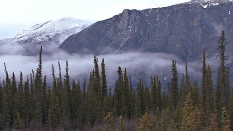 Low clouds and heavy mist creeping over the tundra/taiga/boreal forest in front of an imposing mountain of rock in the late evening in northern Yukon Territory, Canada.