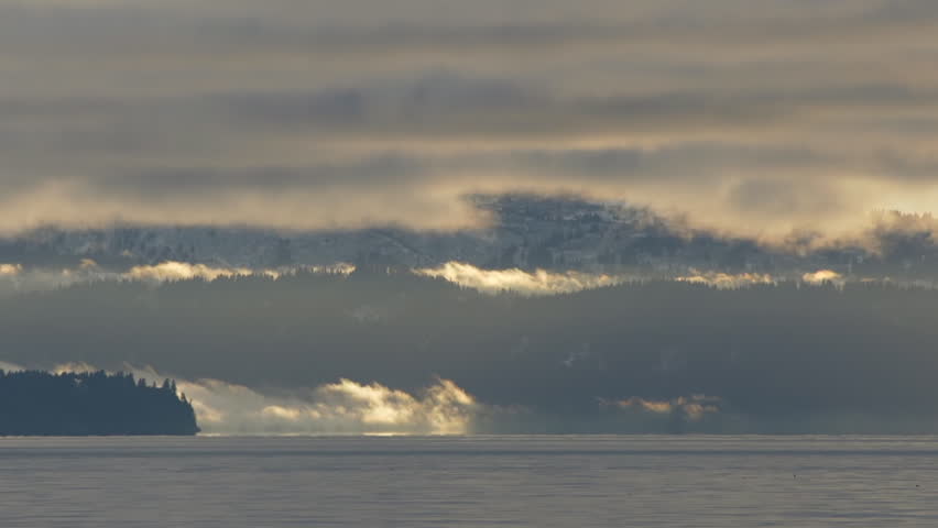 Heavy but luminous low clouds partially obscuring mountains with wispy fog tendrils moving over the water of the bay and the islands in Kachemak Bay, late afternoon, early winter. Time lapse.