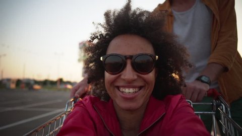 african american girl with curly hair making selfie video while caucasian bearded man circling her in shopping trolley in mall parking lot at sunset slow motion