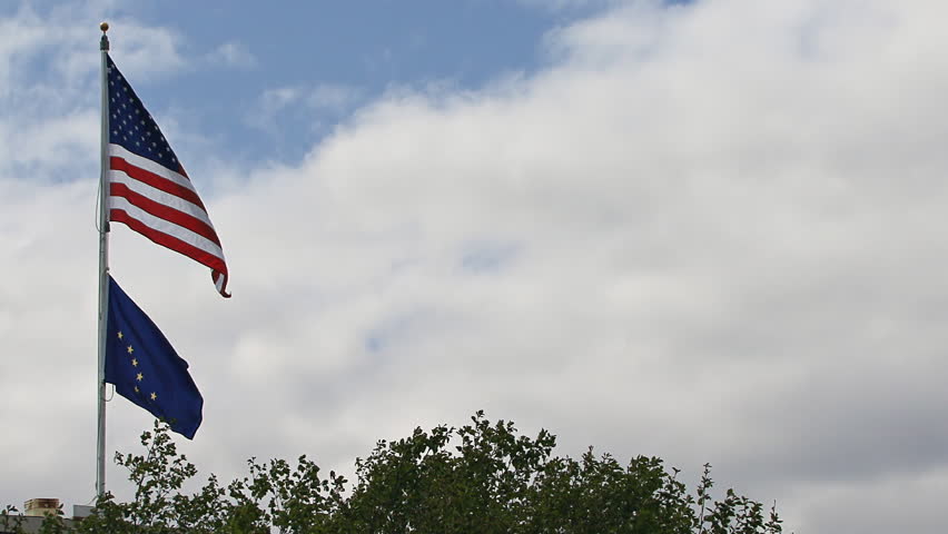 United States flag flying on a windy day over the Alaskan State flag with clouds