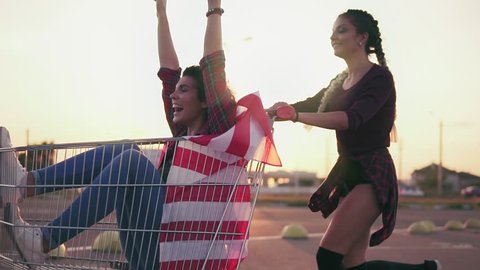 Young hipster teen girls having fun at the shopping mall parking, riding in shopping cart holding the american flag. Lens flare. Slowmotion shot