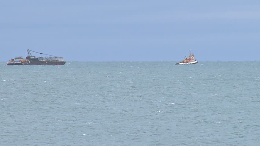 Long shot of a tug boat towing a laden barge chock full of machinery.
