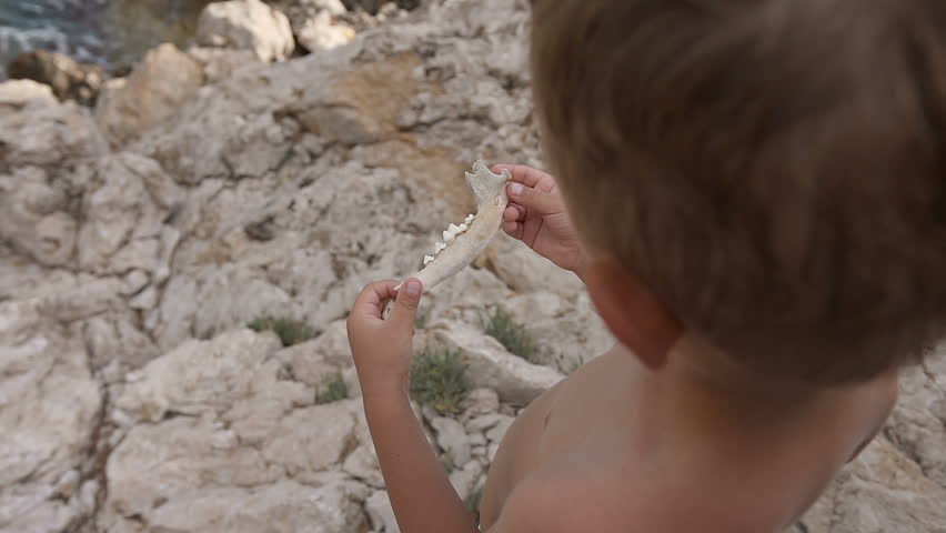 A good boy sits on a cliff by the sea and throws stones in the water. The child on the beach looks at the distance. 50fps