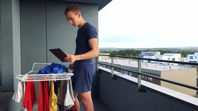 A young man takes down clothes from the drying rack on the balcony in modern apartment and video calls with someone on the tablet 