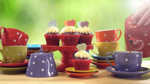 4k Colorful Mad Hatter style tea party with cupcakes and rainbow colored polka dot cups and saucers, with bokeh garden background and lens flare, pouring tea. 
