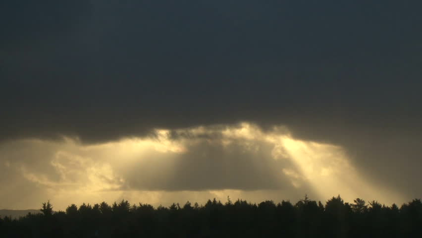Sunlight shining through heavy rain cloud over trees at sunrise, time lapse.