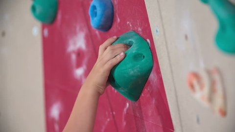 A boy is holding onto the climbing stone when climbing on the climbing wall in the school gym. Climbing stones are of different colors.
