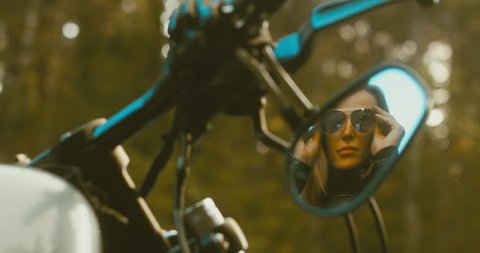 Reflection in rear view mirror, beautiful adult female biker putting on sunglasses before the ride. 4K UHD RAW edited footage