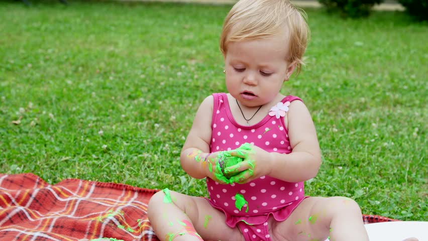 a little child, a one-year-old girl playing, painting with finger paints, decorating herself, in the garden, sitting on a blanket, bedspread, on grass, lawn, in the summer. she