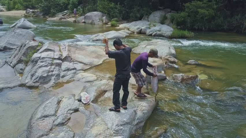 flycam rotates around guy in cap sitting on large stone and taking water into fish bag keeping in river