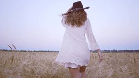Back view of young happy woman in white dress running in the field with hat in hand