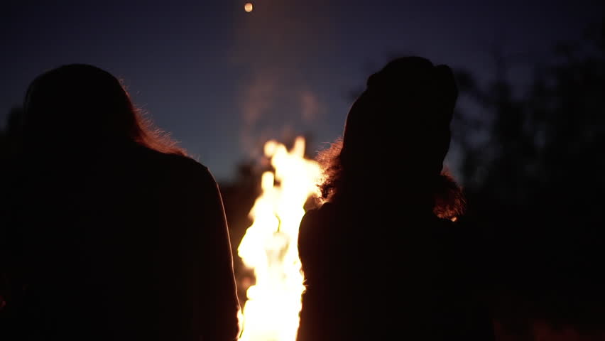 silhouettes of two girls sitting in front bonfire with flame sparks and smoke rising to sky near wood slow motion closeup Royalty-Free Stock Footage #31227322