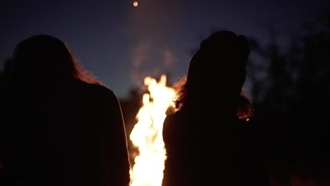 silhouettes of two girls sitting in front bonfire with flame sparks and smoke rising to sky near wood slow motion closeup