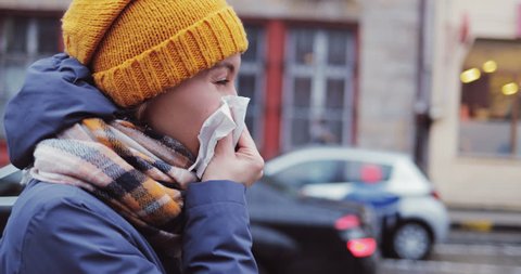Sick Woman Blowing Her Nose Into Tissue Outdoors. SLOW MOTION 4K DCi. Young Female having cold or flu symptoms, standing in a busy city street. Autumn or winter cold weather.