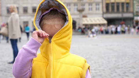 Little Kid Girl Have Fun Playfully Hiding Closing A Zipper Of Yellow Sleeveless Jacket With A Hood