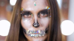 Scary young girl with creative halloween war-paint in the dressing room. Portrait of glamorous skull with rhinestones and sequins. Professional make-up for the celebration. Slow motion.