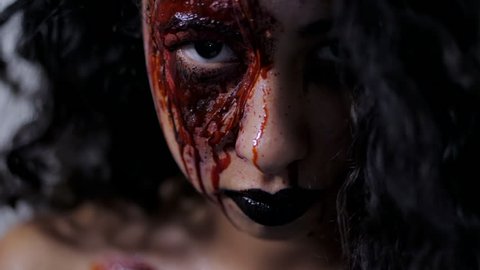 Scary portrait of young girl with Halloween blood makeup. Beautiful latin woman with curly hair looking into camera in studio. Living dead greasepaint. Slow motion.