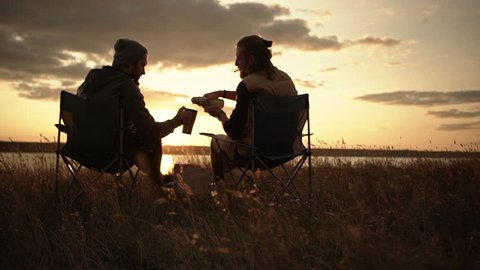 Two men sitting in camping chairs in a field watching sunset pouring tea from thermal mugs.
