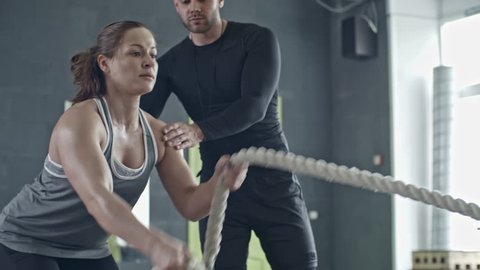 Muscular woman doing battle rope exercise in cross training gym with help of personal coach