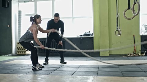Personal coach standing with timer and motivating young woman doing battle rope exercise