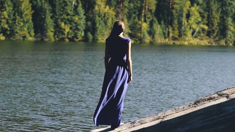Slowmotion of Beautiful young woman in long dress standing on shore near lake