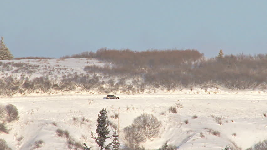 Long shot of SUV driving left to right on snowy road in Alaska.