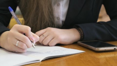 Schoolgirl dressed in a black suit sits at a school desk writing text in exercise book using ballpoint pen next to a golden smart phone. Close-up